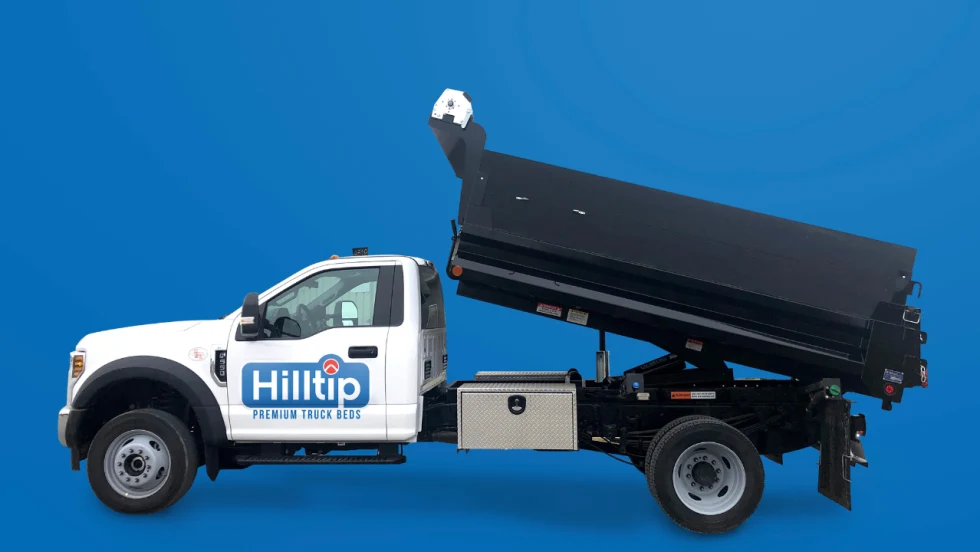 White Hilltip Premium Truck Beds chassis truck with a raised black dump bed, showcasing the hydraulic dumping feature and underbody toolbox against a blue background.