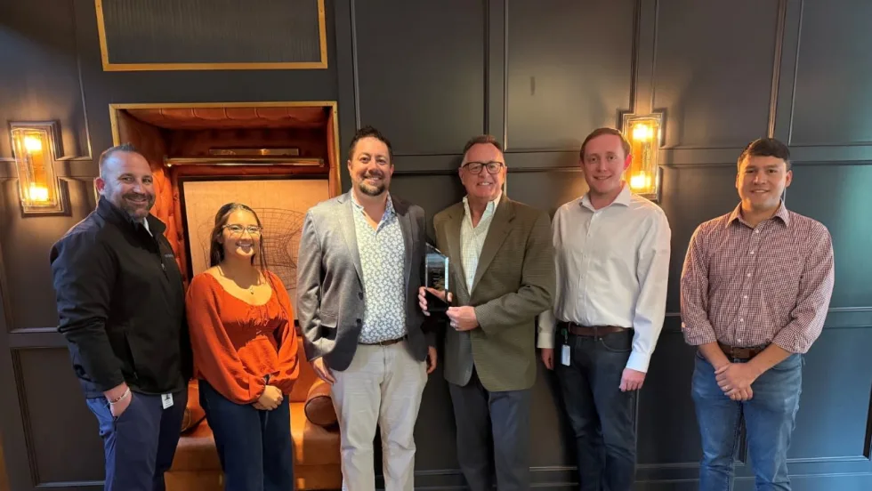 Six employees pose indoors as one team member presents an award to another during a recognition ceremony. The group stands in front of a dark-paneled wall with warm lighting, smiling for the camera.