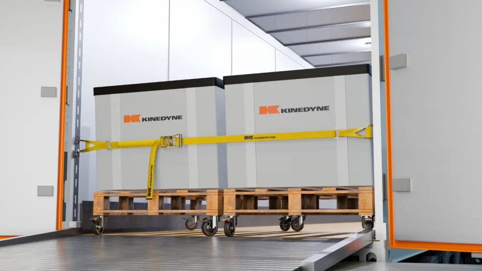 Kinedyne cargo securement strap holds two large containers on wheeled pallets inside a truck trailer, demonstrating a heavy-duty logistics load restraint system.