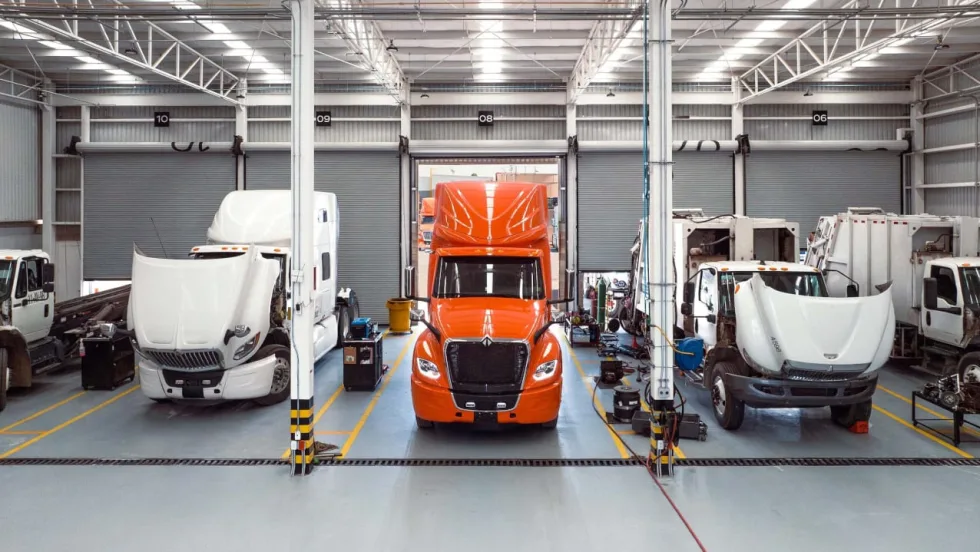 Commercial trucks undergoing maintenance inside a large service facility, including an orange semi-truck centered between white trucks with raised hoods and service equipment.