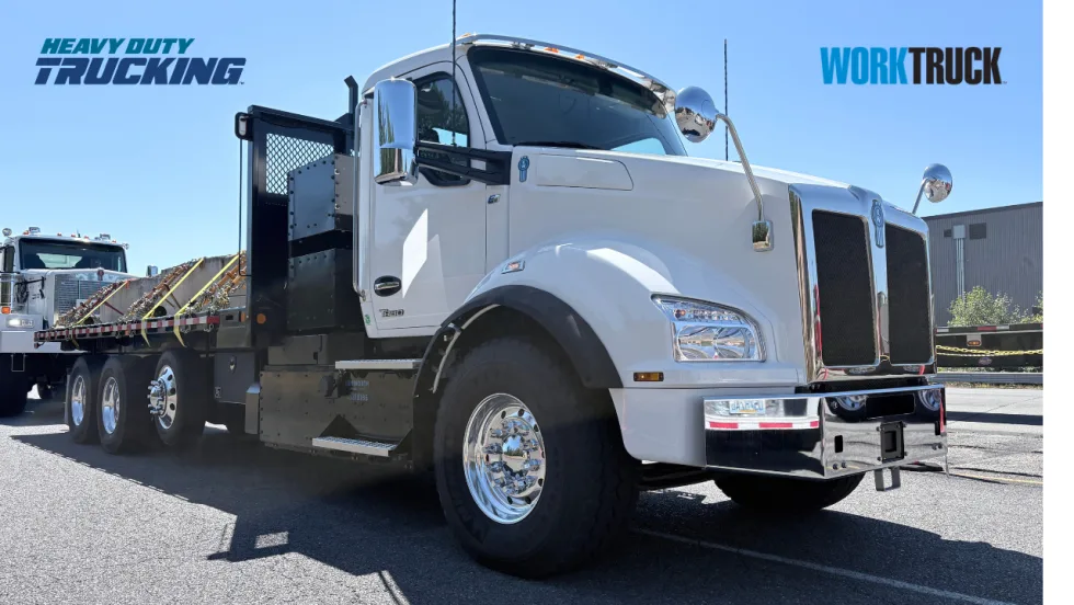 White Kenworth T880 heavy-duty flatbed truck displayed outdoors, featuring chrome accents and rugged tires, with another work truck visible in the background.