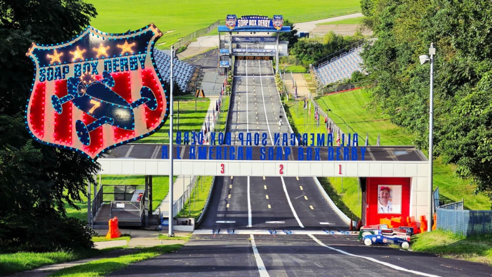 View down the 996-foot Soap Box Derby track at Derby Downs in Akron, Ohio, with grandstands, the starting structure, and a colorful Soap Box Derby sign in the foreground.