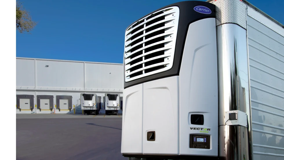 Carrier Transicold Vector 8200 refrigeration unit mounted on a refrigerated trailer outside a distribution facility with loading docks and additional refrigerated trucks.
