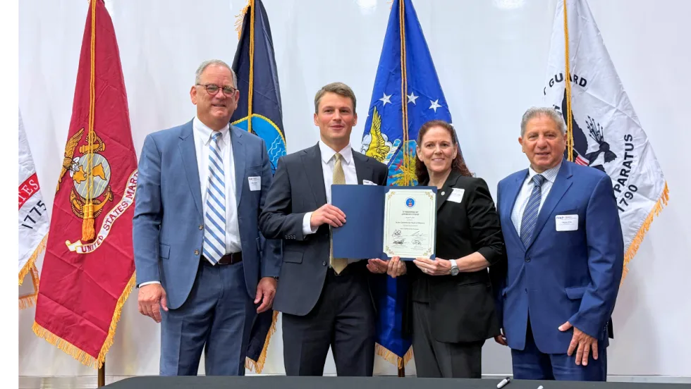 Four representatives pose with a signed certificate during an Isuzu and IVAP event, standing in front of U.S. military service flags.