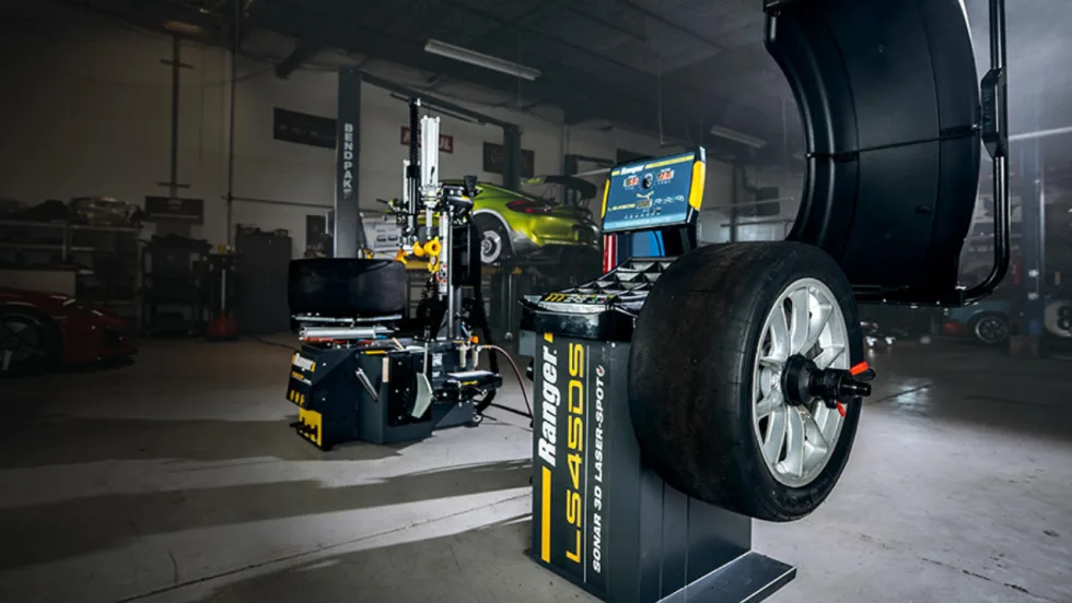 Ranger tire changer and wheel balancer equipment displayed in an automotive service shop, with a mounted performance tire and vehicles on lifts in the background.