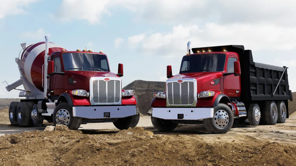 Two red Peterbilt vocational trucks, a concrete mixer and dump truck, parked at a construction site with dirt piles and a partly cloudy sky in the background.