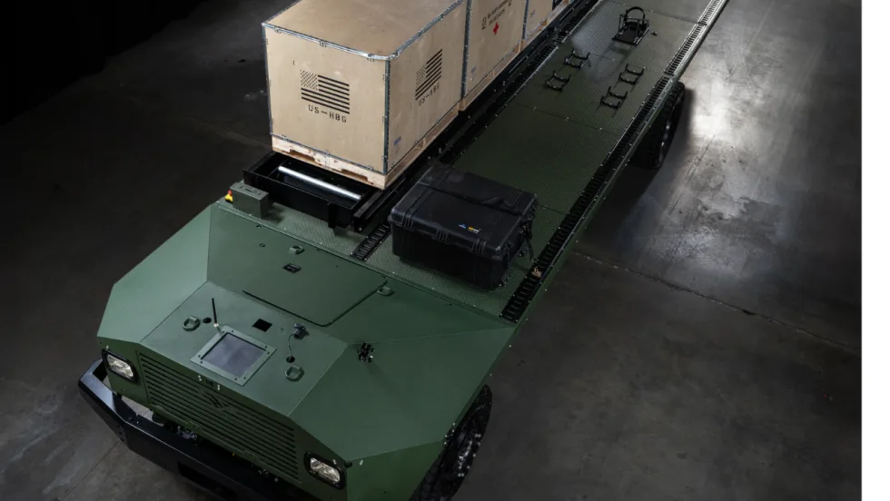 Green military cargo vehicle carrying large shipping crates and equipment on its flatbed, photographed from above inside an industrial facility.