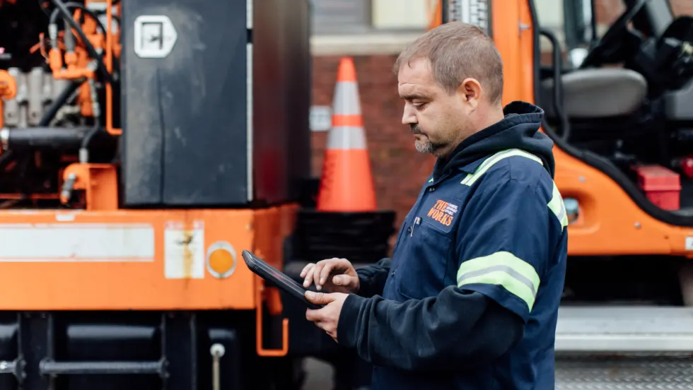 Fleet maintenance worker using a tablet beside an orange work truck and service equipment.