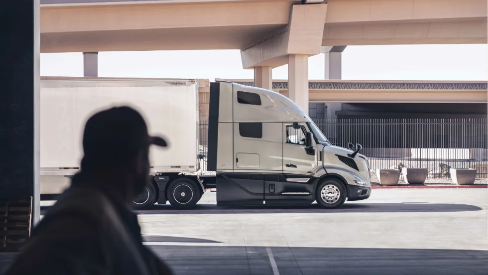 Volvo semi-truck with a white box trailer parked beneath highway overpasses, viewed from a loading area with a silhouetted person in the foreground.