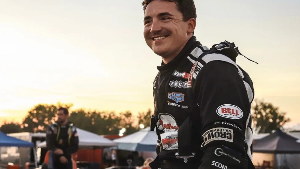 Isaac Johnson in a black-and-white firesuit smiles at the track at sunset with tents, equipment, and another person blurred in the background.
