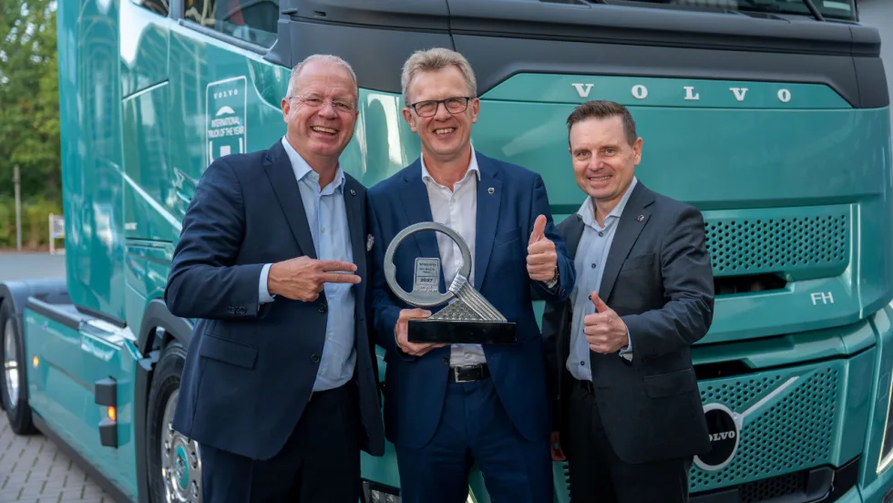 Three Volvo executives pose with a 2027 International Truck of the Year trophy in front of a teal Volvo FH truck, with two giving thumbs-up gestures.
