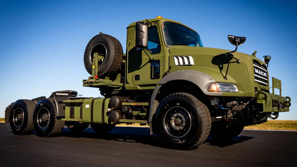 Olive-drab Mack Defense military tactical truck chassis with rugged tires, spare tire mounted behind the cab, heavy-duty front bumper, and three-axle configuration, photographed under a clear blue sky.