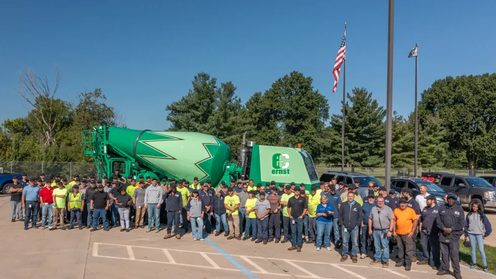Large group of employees gathered outdoors in front of a green Ernst Concrete mixer truck, posing beneath American and company flags to commemorate a company milestone.