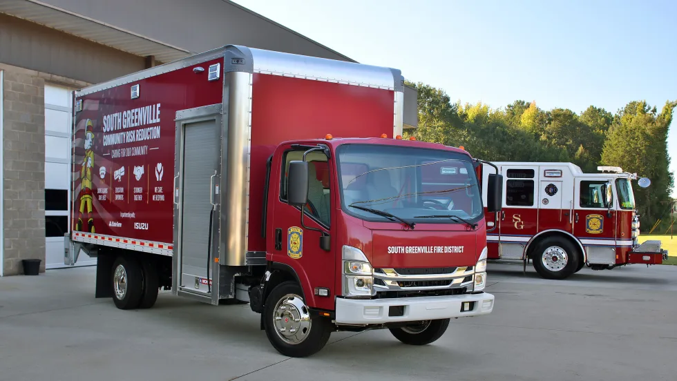 South Greenville Fire District red Isuzu box truck featuring Community Risk Reduction graphics, parked outside a fire station with a red-and-white fire engine in the background.
