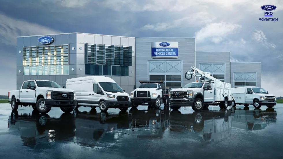 Ford commercial trucks and vans, including pickups, a Transit cargo van, a medium-duty truck, and a utility truck, parked outside a Ford Commercial Vehicle Center.