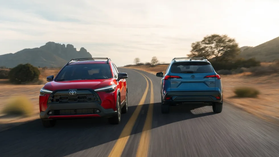 Red and blue 2027 Toyota Corolla Cross SUVs driving side by side on a two-lane road.