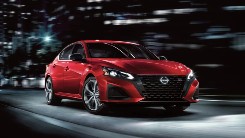 Red 2027 Nissan Altima SR sedan driving through a city at night, shown from the front three-quarter angle with illuminated headlights and dark alloy wheels.