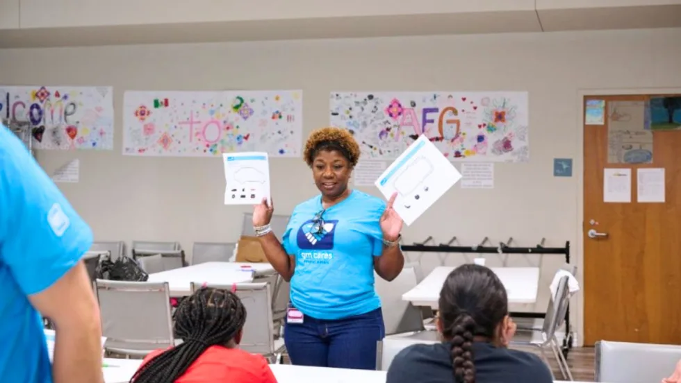 A woman wearing a blue GM Cares shirt holds up educational materials while speaking to a group of participants seated at tables during a community outreach or educational workshop.