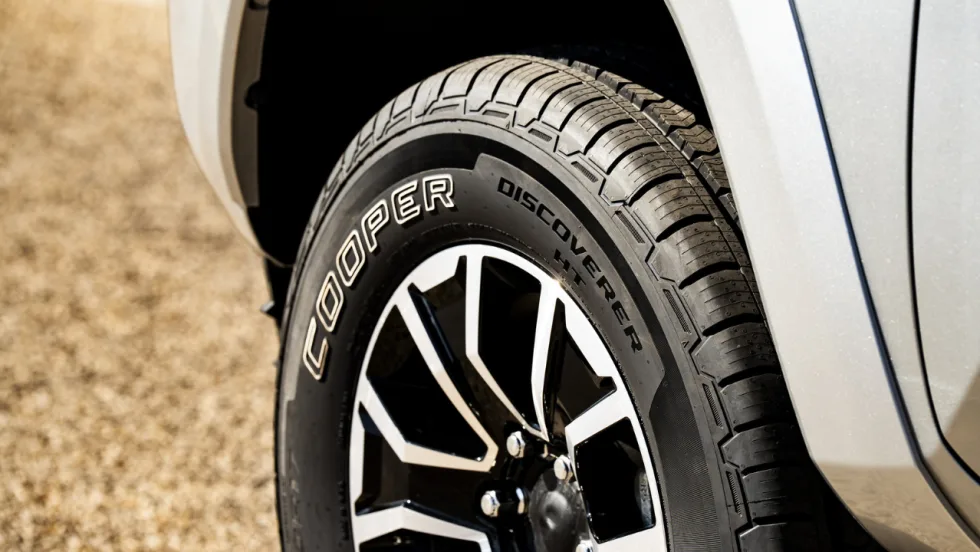 Close-up of a Cooper Discoverer HT tire mounted on a black-and-silver alloy wheel of a silver pickup truck, highlighting the tire’s tread pattern and Cooper branding on the sidewall.