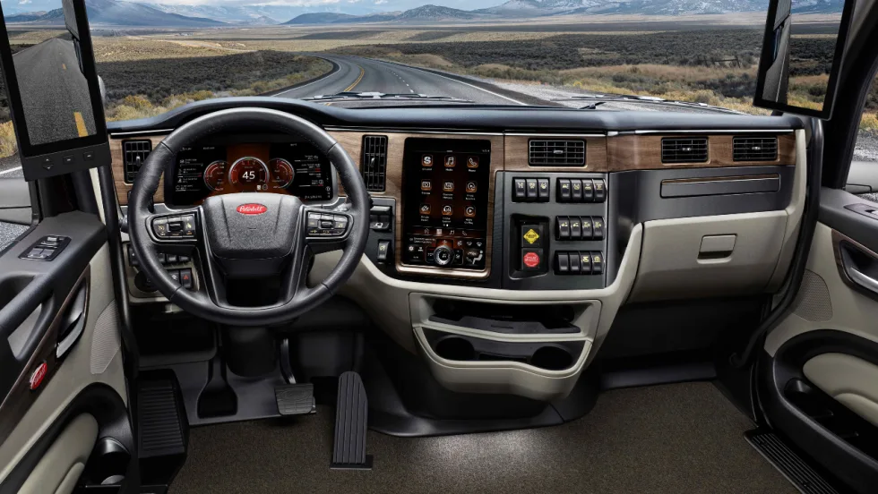Interior view of a Peterbilt commercial truck cab featuring a digital instrument cluster, touchscreen infotainment system, steering wheel, dashboard controls, and digital side mirrors, with a winding highway and mountains visible through the windshield.