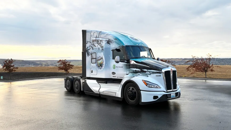 A white Kenworth sleeper cab truck featuring Boise Cascade branding and winter-themed graphics is parked on wet pavement overlooking rolling hills beneath a cloudy sky.