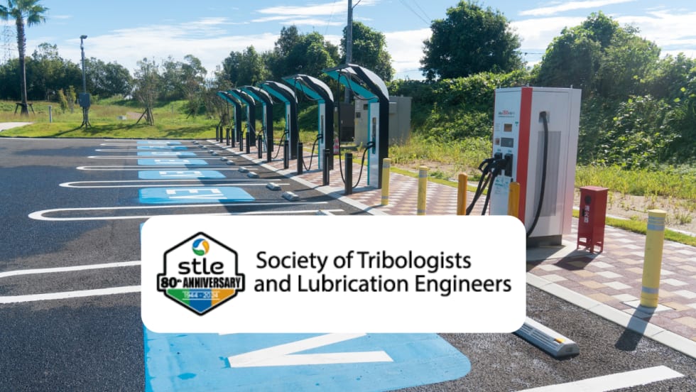 Electric vehicle charging station with multiple chargers and designated EV parking spaces, overlaid with the Society of Tribologists and Lubrication Engineers (STLE) 80th-anniversary logo.