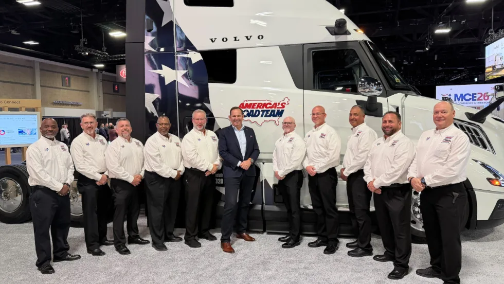Ten members of the America's Road Team pose in front of a white Volvo semi-truck featuring patriotic graphics and the America's Road Team logo at the MCE26 conference and exhibition.