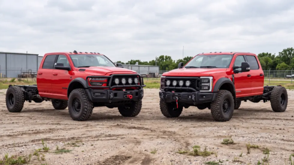 Red Ram and Ford crew cab chassis trucks parked on a gravel lot, with oversized off-road tires, front winches, auxiliary lights, and exposed rear frame rails.