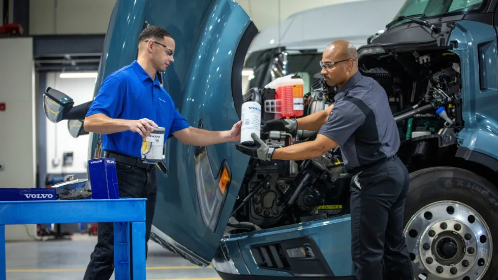 Two Volvo truck service technicians wearing safety glasses exchange maintenance filters beside a blue Volvo semi-truck with its hood raised, exposing the engine, inside a commercial vehicle service facility.