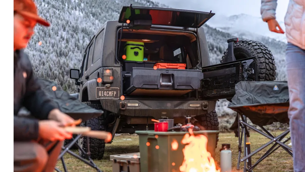 Rear view of a Jeep Wrangler at a mountain campsite, featuring an open tailgate and DECKED storage drawer system loaded with camping gear, while campers sit around a fire in a snowy landscape.