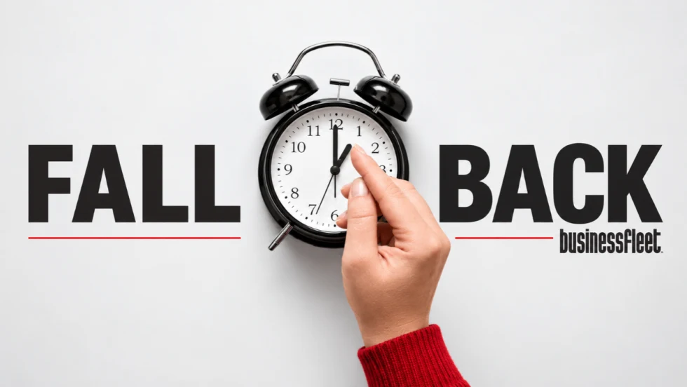Hand in a red sweater adjusts a black alarm clock between the words “Fall Back,” with the Business Fleet logo on a light gray background.