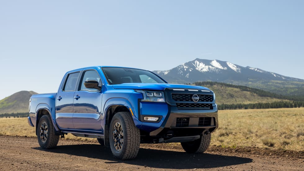 Blue 2027 Nissan Frontier Sport Edition pickup truck parked on a dirt road with snow-capped mountains in the background, showcasing its off-road styling and all-terrain tires.