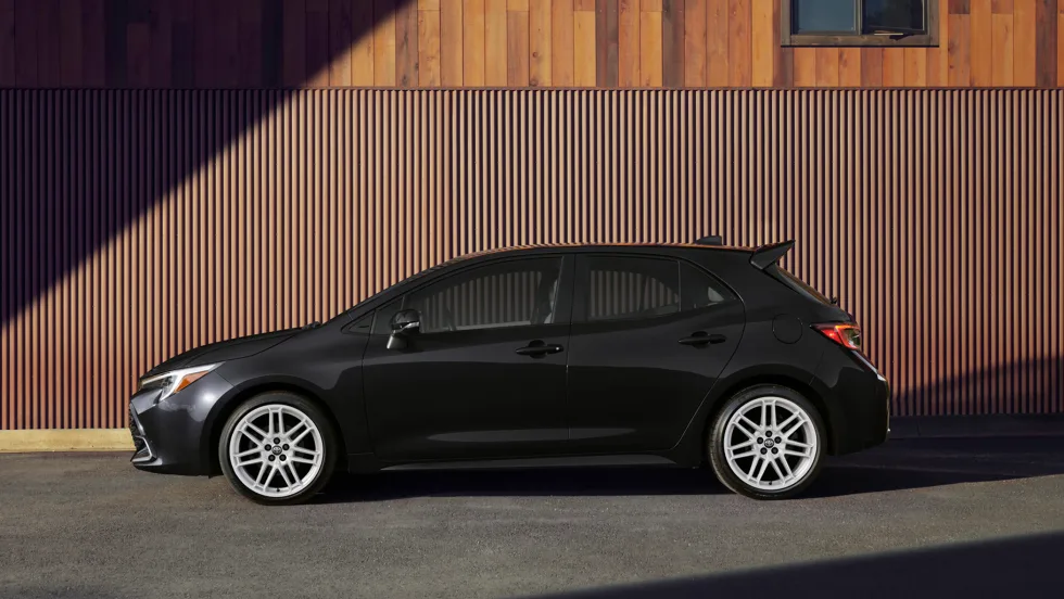 Side profile of a black 2027 Toyota Corolla Hatchback parked in front of a wood-paneled building, showing the compact hatchback's available 18-inch gloss white alloy wheels and updated styling.