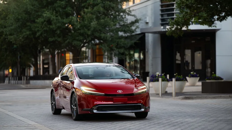 Front three-quarter view of a red 2027 Toyota Prius parked in an urban setting, showcasing the updated hybrid sedan's sleek design, LED lighting and aerodynamic styling.