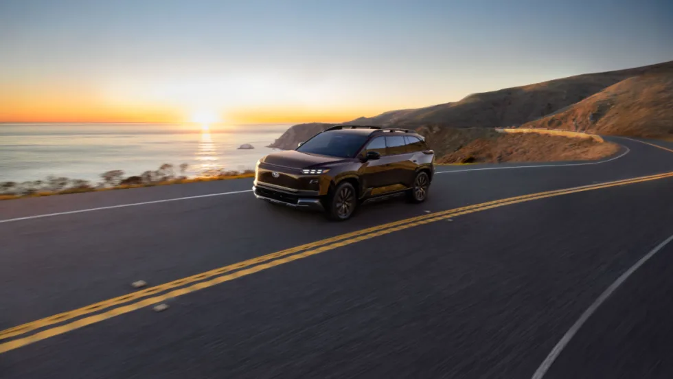 Subaru electric SUV driving along a coastal highway at sunset, representing the 2027 Getaway with three-row seating and all-electric performance.