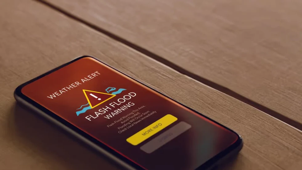 Mobile phone displaying a flash flood weather alert on screen illustrating real-time weather warnings powered by GPS and cloud processing for field safety.