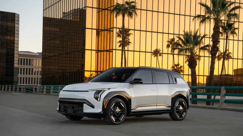 White Kia EV3 electric SUV parked in an urban setting at sunset with palm trees and glass buildings in the background.