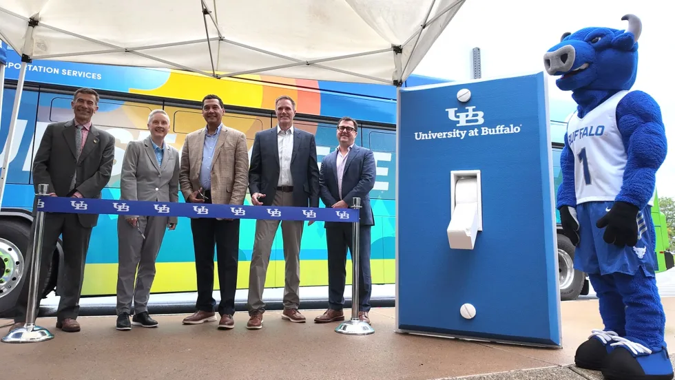 Five University at Buffalo representatives stand behind a ribbon beside a colorful transit bus and UB-branded charging station, with the Victor E. Bull mascot nearby.
