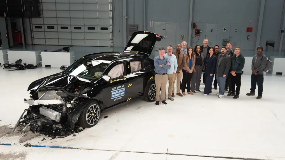 A group of people stand next to a crashed car for IIHS testing