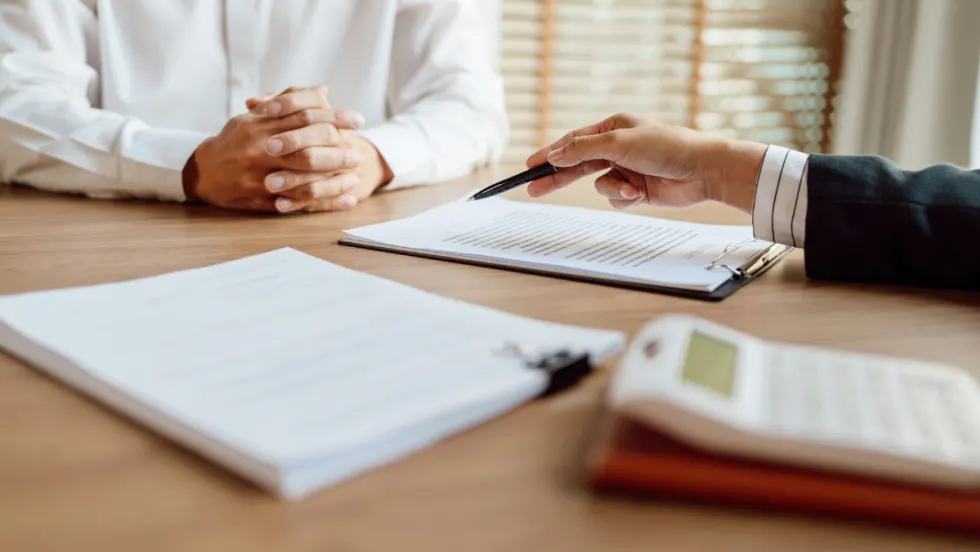 Two people sit across from each other at a desk during a business meeting. One person, wearing a white shirt, has their hands folded while the other gestures with a pen toward documents clipped to a clipboard. Additional paperwork and a calculator are visible on the table, suggesting a discussion involving contracts, finances, or administrative paperwork. Sunlight filters through window blinds in the background, creating a professional office setting.