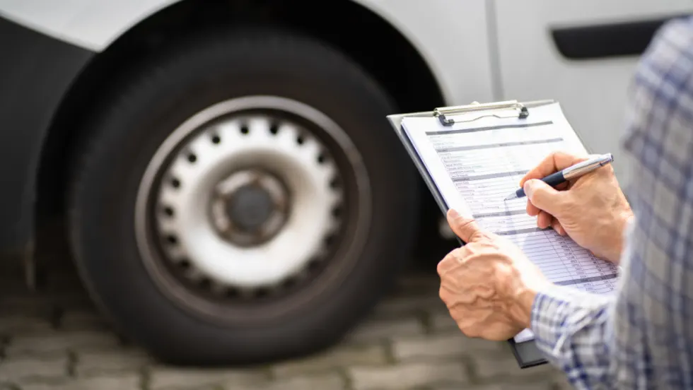 A person holding a clipboard and writing on an inspection checklist beside the wheel of a large white vehicle, likely conducting a fleet or safety inspection.