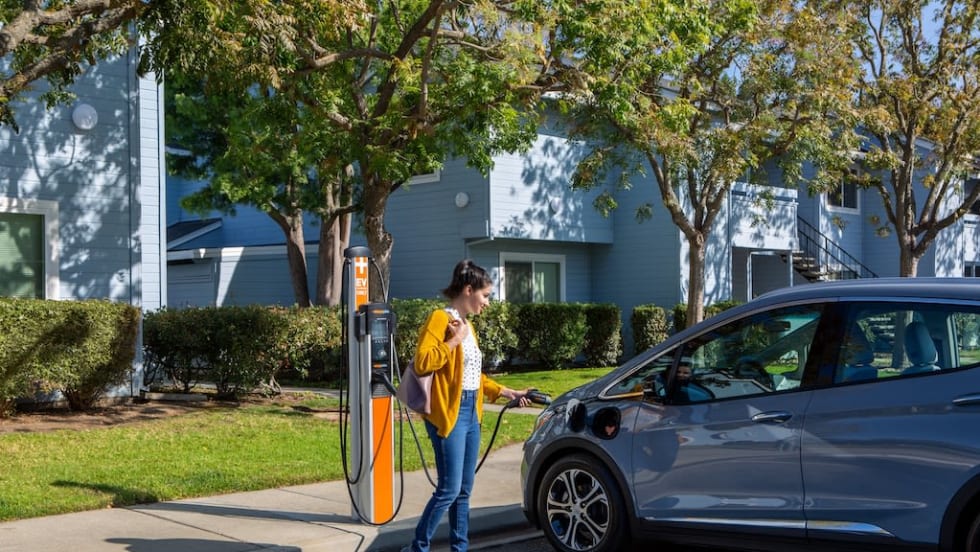 Woman plugs an electric vehicle into a ChargePoint charger outside an apartment complex, representing expanded EV charging access for multifamily housing.