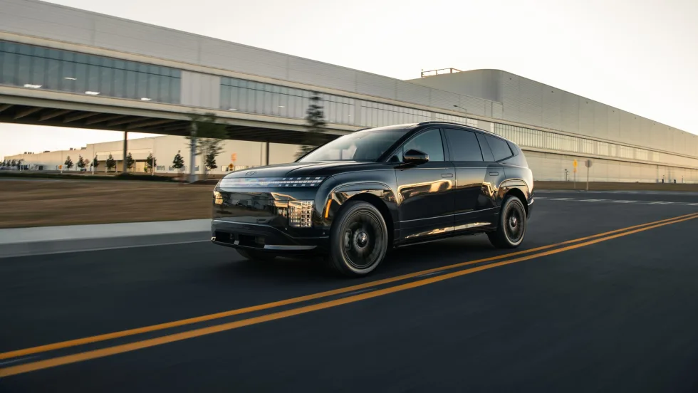 Black Hyundai IONIQ 9 electric SUV driving on a road near a modern industrial building.