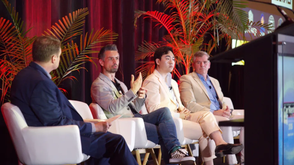 Moderator and three panelists on stage in white chairs in front of a red curtain and palm frond planters.