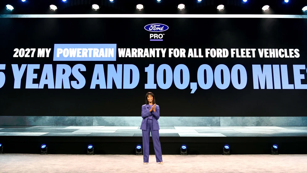 Presenter stands onstage before a large screen announcing Ford Pro’s expanded 5-year, 100,000-mile powertrain warranty for additional fleet vehicles beginning with the 2027 model year.