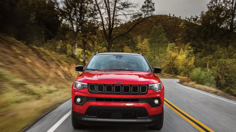 A red 2027 jeep compass drives on the road surrounded by foliage