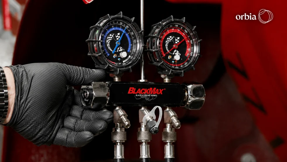 A gloved hand holds a gauge manifold with blue and red pressure gauges and three connected hoses.