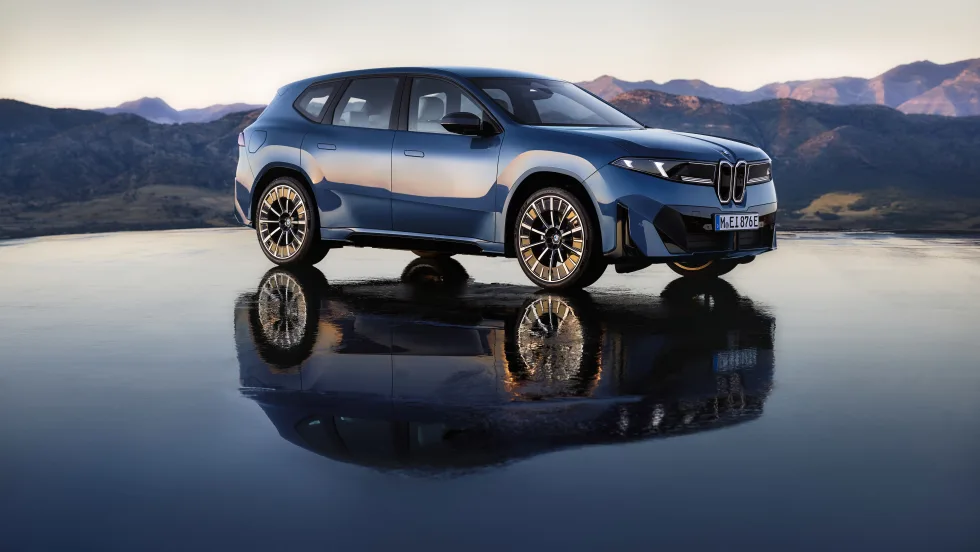 Blue BMW iX3 electric SUV parked on a reflective surface with mountain scenery in the background at dusk.