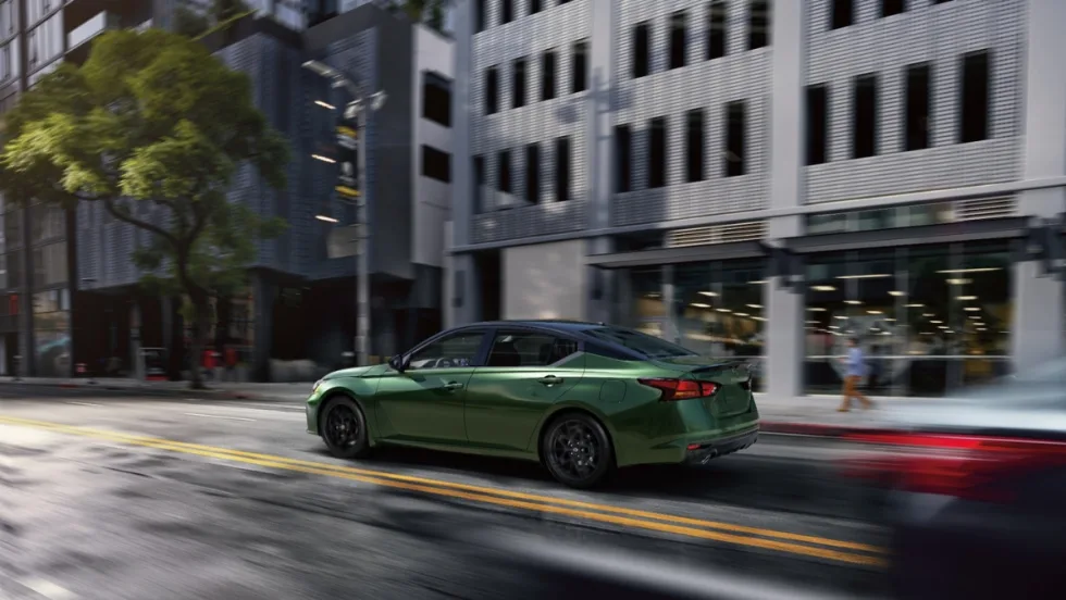 The 2026 Nissan Altima drives on a road between white buildings. 