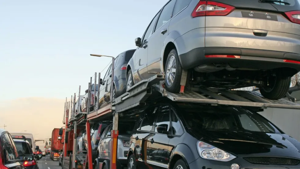 A double-decker stinger transport trailer loaded with cars drives down a crowded freeway. 